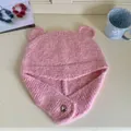 Detailed view of pink knitted bear-ear beanie with silver heart button on cream desk - Pink Heather