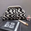 Close-up of a stylish wave-patterned knitted clutch bag alongside elegant lifestyle props. - Black Beige