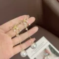 Detail of hand holding long golden bow earrings with elegant dangling pearls and zircons. - Bow Pearl Drop Style (E3142) / Gold White