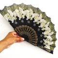 Detalle de un abanico plegable negro con motivos de mariposas doradas sostenido a mano - Flor Mariposa - Amarillo