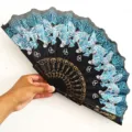 Vista detallada de un abanico negro con mariposas azules brillantes y varillas doradas. - Flor Mariposa - Azul