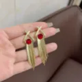 Detail of gold tassel drop earrings with red crystal accents held for size comparison - Hollow Hoop Tassel Style (E3596) / Gold Red