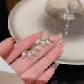Primo piano di eleganti orecchini a lobo con perle e base dorata tenuti su dita curate. - Stile Perla a Goccia a Ventaglio (E3113) / Oro
