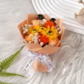 Close-up detail of artificial flower bouquet with sunflowers and roses in orange wrap. - Champagne Roses and 2 Sunflowers with Dried Flowers