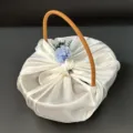 Detail view of a woven gift basket wrapped in white satin with blue floral accents. - Basket + Wrap + Flowers / White
