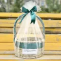 Detailed view of a round woven straw gift basket wrapped in tulle with a dark green ribbon bow. - Basket With Cream Mesh And Ribbon / Vintage Green