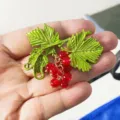 Close-up of a vintage baroque grape brooch with green leaves and red beads held in hand. - Red