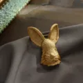 Detailopname van een vintage matgouden konijnenkopbroche op grijze zijde. - Vintage Konijnenkop (Stijl 2) / Mat Goud