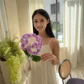 Detailed view of a handmade purple crocheted carnation flower held by a smiling model - Large Single Carnation (Purple) / Single Stem Without Bag / Purple