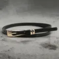 Close-up of black leather thin belt with gold metal buckle detail on gray texture - Black