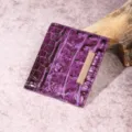 Vista detallada de un tarjetero morado con textura de cocodrilo y adorno metálico dorado. - Tarjetero (PG832) / Púrpura profundo