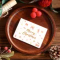 Gold foil Merry Christmas greeting card displayed on a rustic wooden tray with festive props - Bell Star Style