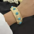Klassischer goldfarbener breiter Armreif mit türkisfarbenen Schmucksteinen am Handgelenk - Blauer Türkis