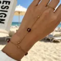 Detailed view of a gold hand chain featuring red flower charms and beads on a beach. - Purple Artificial Pearl Hand Chain
