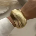 Makroansicht eines breiten, goldenen Metallarmreifs mit Textur zu einer weißen Seidenbluse. - Breiter Gestreifter Gebogener Armreif (217) / Gold