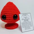 Close-up of a red crochet emotional support plush with a humorous greeting card - Red Plug With Large Card
