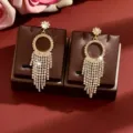 Close-up of gold-toned rhinestone tassel earrings with geometric circle and floral stud. - Gold