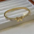 Eleganter goldfarbener Titanstahl-Armreif mit Schleife und Kristallakzenten auf Tablett. - Großer Schleifen Strass Stil / 4mm Breit / Gold