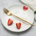 Hochwertige goldene Edelstahl-Gabel auf weißem Keramikteller mit frischen Erdbeeren. - Speisegabel / Gold