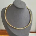 Primer plano de un elegante collar gargantilla de metal dorado en un soporte gris. - Ajustable / Dorado