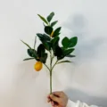 Primo piano di una mano che tiene un ramo di limone artificiale con frutti gialli - Ramo di Limone / 1 Pezzo
