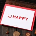 Λευκή ευχετήρια κάρτα με σχέδιο 'HAPPY' και κόκκινο φάκελο σε ξύλινη επιφάνεια. - Σχέδιο Happy Χαμόγελο