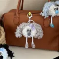 Close-up of a cute curly grey plush doll keychain hanging on a brown leather bag. - Gray
