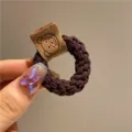 Vue détaillée d'un chouchou tressé marron avec une étiquette en tissu ourson mignon. - Brun Foncé