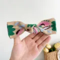 Détail d'un accessoire pour cheveux avec nœud brodé de style bohème tenu à la main devant un mur - Petite Pince Nœud Fleurie Brodée / Kaki