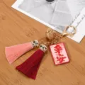 Gros plan sur l'étiquette rouge et les pompons bicolores de ce porte-clés doré à clochette - Paix (Rose + Gland Rouge)