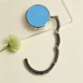 Detailed view of a portable foldable bag hook with a blue finish on a minimalist desk - Folding Hook / Blue