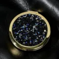 Detailed close-up of circular gold compact mirror with sparkling black and blue glitter. - 70mm / Obsidian color (gold frame)