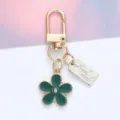 Close-up of a green daisy charm and engraved 'Hand Made' tag on a gold metal keychain. - Daisy Pendant (With Hand Made Tag) / Dark Green