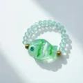 Macro view of a handcrafted green glass fish charm on a blue beaded elastic ring. - Clear