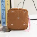 Close-up view of brown quilted cosmetic bag featuring white bow embroidery and pink pull. - Brown
