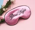 Detailansicht einer rosa Satin-Schlafmaske mit schwarzem Good Night-Stickerei-Text. - Rosa