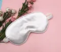 Detailed view of a soft white satin sleep mask placed on a pink floral background. - Pearl White