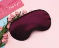 Close-up of a burgundy satin sleep eye mask with black piping on a pink backdrop - Purple
