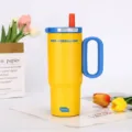 Detail view of yellow insulated tumbler with blue handle and straw, set in a clean floral scene. - Yellow