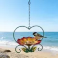 Heart-shaped metal bird feeder with flower tray hanging over a scenic beach landscape. - Red