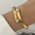 Close-up van een elegante gedraaide gouden armband met twee parelaccenten om een pols. - Goud (Onregelmatig Geometrisch Dubbele Parel Open)