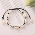 A detailed macro view of a bohemian woven cowrie shell bracelet lying on a book background. - Black Braided Rope with Creamy White Shell (BR23Y0237-2)