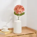 Detailed view of artificial hydrangea flower in white ceramic vase on wooden desktop setup. - Orange