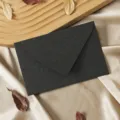 Close-up detail of a textured black envelope on a satin fabric with wooden tray and dry leaves. - Cashmere Texture Charcoal Black Envelope, 1 Sheet