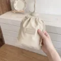 Detailopname van een minimalistisch beige opbergzakje met trekkoord en een FHUASHIJIE-patch. - S / Beige