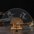 Élégant éventail pliant chinois traditionnel en tissu noir, baleines en bois et support. - N° 2 Pureté