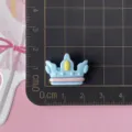 Close-up of a glossy blue crown resin accessory with pink and yellow details on a grid mat. - Blue
