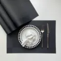 Detailed view of black woven PVC placemat set with elegant tableware on neutral tabletop. - Black Single Frame