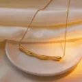 Close-up detail of a minimalist gold irregular curved bar pendant necklace on a sleek ceramic plate. - Pleated Style / Gold