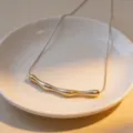 Detailed view of a stylish irregular curved metal arc tube necklace placed in a white plate. - Pleated Style / Steel